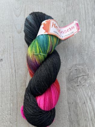Ein Strang handgefärbter Wolle liegt auf hellem, leicht strukturiertem Holz. Die Wolle zeigt kräftige Farbabschnitte: tiefes Schwarz, leuchtendes Pink, sattes Rot, strahlendes Türkis, frisches Grün, Gelb und violette Übergänge. Die Banderole trägt das Her